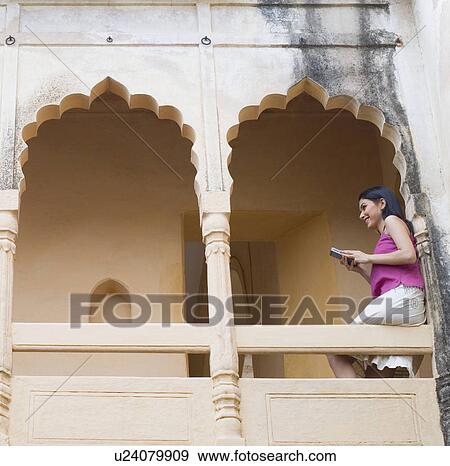 Low angle view of a young woman sitting on the railing of a palace and operating a mobile phone, Neemrana Fort Palace, Neemrana, Alwar, Rajasthan, India View Large Photo Image Stock Photo - Low angle view of a young woman sitting on the railing of a palace and operating a mobile phone, Neemrana Fort Palace, Neemrana, Alwar, Rajasthan, India. Fotosearch