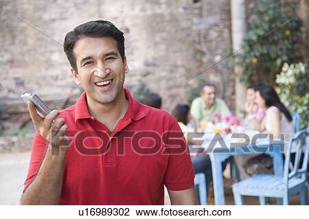 Stock Image - Portrait of a mid adult man holding a mobile phone and smiling with his friends sitting in the background. Fotosearch