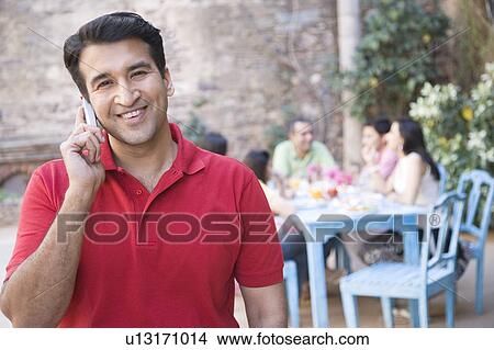 Portrait of a mid adult man talking on a mobile phone with his friends sitting at a dining table in the background View Large Photo Image Picture - Portrait of a mid adult man talking on a mobile phone with his friends sitting at a dining table in the background. Fotosearch