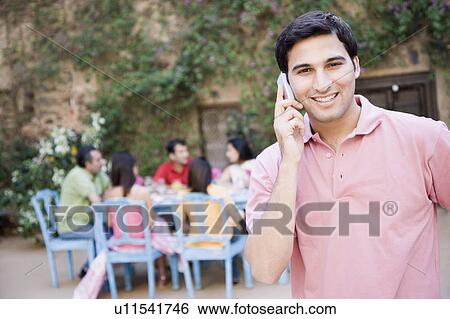 Portrait of a young man talking on a mobile phone and his friends sitting at a dining table in the background View Large Photo Image Stock Photograph - Portrait of a young man talking on a mobile phone and his friends sitting at a dining table in the background. Fotosearch