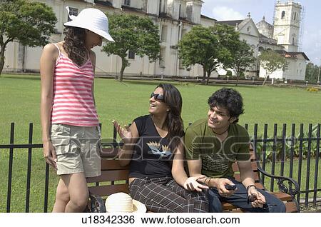 Young couple sitting on a bench with another young woman standing beside them, Se Cathedral, Goa, India View Large Photo Image Stock Photograph - Young couple sitting on a bench with another young woman standing beside them, Se Cathedral, Goa, India. Fotosearch