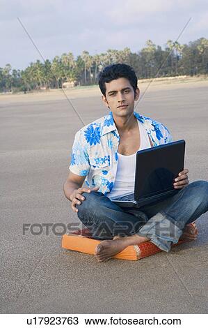 Stock Image - Portrait of a young man sitting on a cushion and holding a laptop. Fotosearch