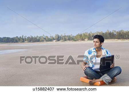 Young man sitting on a cushion and holding a laptop View Large Photo Image Stock Photo - Young man sitting on a cushion and holding a laptop. Fotosearch