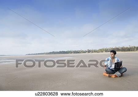 Stock Photo - Young man sitting on the beach and using a laptop. Fotosearch