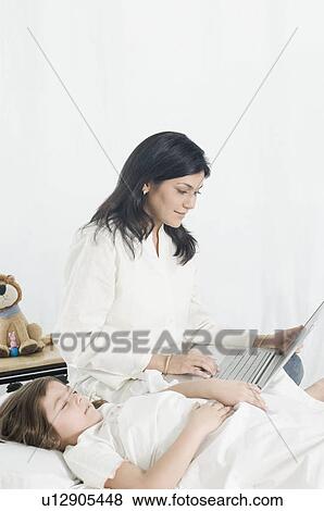 Stock Photo - Mid adult woman using a laptop beside her daughter lying on the bed. Fotosearch