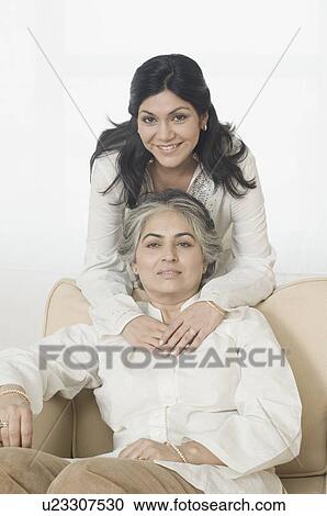 Stock Image - Portrait of a mature woman sitting on a couch and her daughter embracing her from behind. Fotosearch