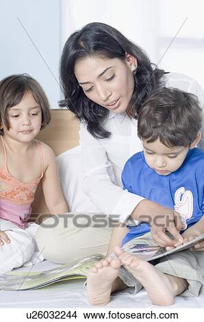 Young woman showing a magazine to her children on the bed View Large Photo Image Picture - Young woman showing a magazine to her children on the bed. Fotosearch
