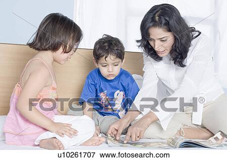 Stock Photo - Young woman showing a magazine to her children on the bed. Fotosearch