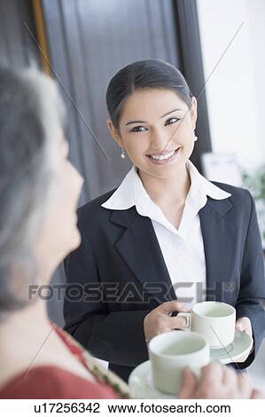 Stock Image - Close-up of two businesswomen holding cups of tea and smiling. Fotosearch