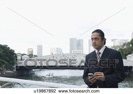 Businessman leaning against a railing and holding a palmtop, Singapore View Large Photo Image Stock Image - Businessman leaning against a railing and holding a palmtop, Singapore. Fotosearch