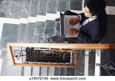 Stock Photography - High angle view of a businessman sitting on the staircase and using a laptop. Fotosearch