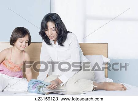 Stock Image - Young woman reading a book with her daughter on the bed. Fotosearch