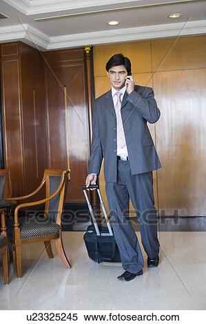 Businessman pulling his luggage and talking on a mobile phone in a hotel View Large Photo Image Stock Photography - Businessman pulling his luggage and talking on a mobile phone in a hotel. Fotosearch