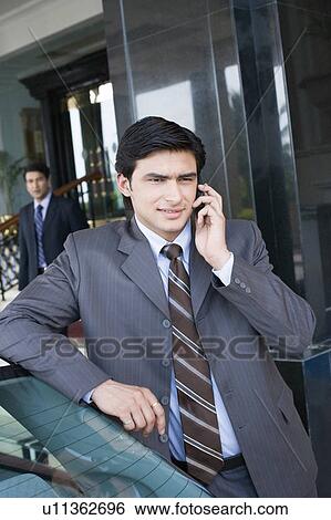 Businessman talking on a mobile phone with another businessman standing in the background View Large Photo Image Stock Photograph - Businessman talking on a mobile phone with another businessman standing in the background. Fotosearch
