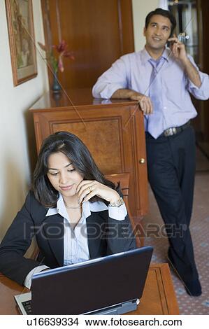 Businesswoman working on a laptop with a businessman talking on a mobile phone in the background View Large Photo Image Picture - Businesswoman working on a laptop with a businessman talking on a mobile phone in the background. Fotosearch