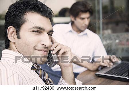 Side profile of a businessman talking on a mobile phone with another businessman using a laptop in the background View Large Photo Image Stock Photograph - Side profile of a businessman talking on a mobile phone with another businessman using a laptop in the background. Fotosearch