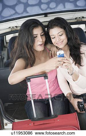Stock Photograph - Two young women sitting in a car trunk and looking at a mobile phone. Fotosearch