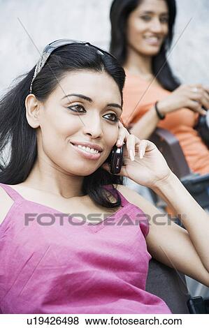 Young woman talking on a mobile phone with another young woman smiling behind her View Large Photo Image Stock Photo - Young woman talking on a mobile phone with another young woman smiling behind her. Fotosearch