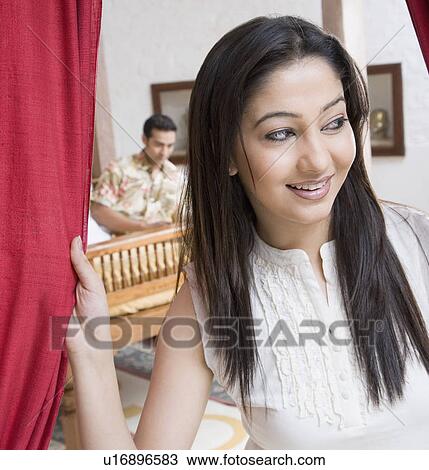 Stock Image - Close-up of a young woman holding curtains with a young man lying in the background. Fotosearch
