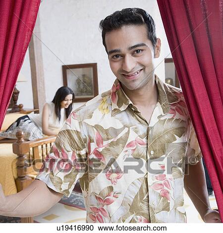 Stock Image - Portrait of a young man smiling with a young woman kneeling on the bed in the background. Fotosearch