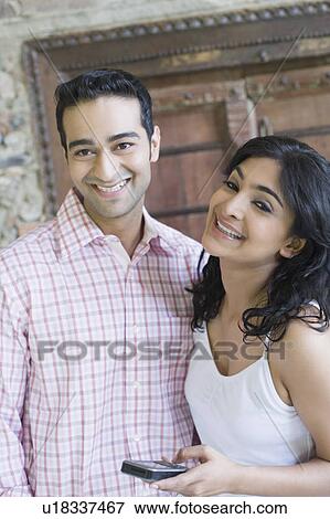 Stock Photo - Portrait of a young woman holding a mobile phone with a young man standing beside her. Fotosearch