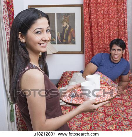Portrait of a young woman holding coffee cups in a serving tray with a young man reclining on the bed View Large Photo Image Stock Photo - Portrait of a young woman holding coffee cups in a serving tray with a young man reclining on the bed. Fotosearch