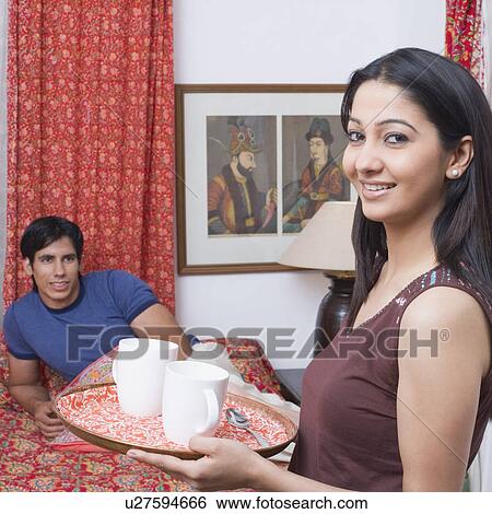 Stock Photograph - Portrait of a young woman holding coffee cups in a serving tray with a young man reclining on the bed. Fotosearch