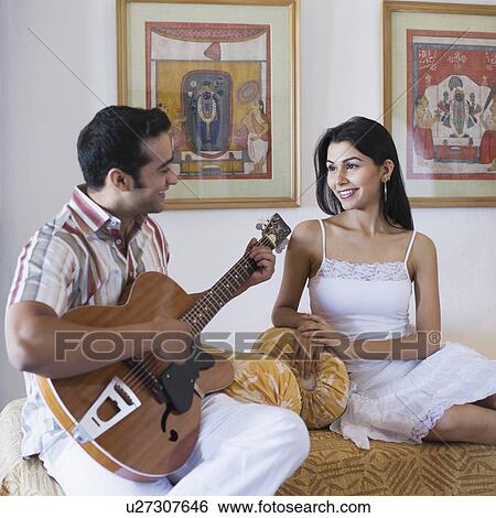 Stock Photograph - Side profile of a young man sitting with a young woman and playing a guitar. Fotosearch