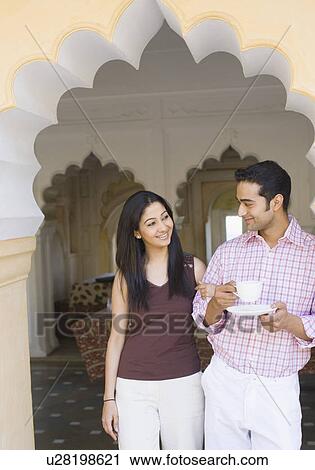 Stock Image - Young woman standing arm in arm with a young man holding a cup of tea. Fotosearch