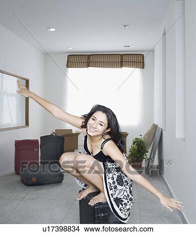 Picture - Portrait of a young woman sitting with her arms outstretched on a suitcase and smiling. Fotosearch