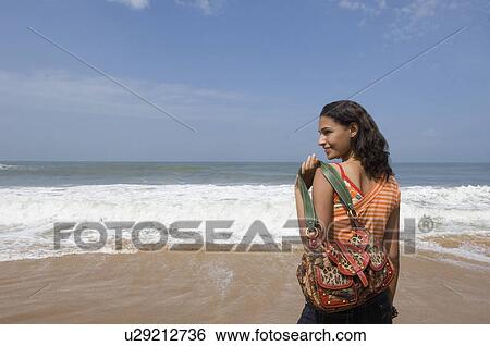 Stock Photograph - Rear view of a young woman carrying a shoulder bag on the beach. Fotosearch