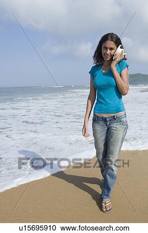Stock Image - Young woman holding a conch shell to her ear and walking on the beach. Fotosearch
