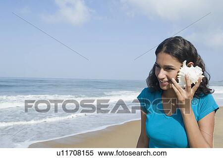 Young woman holding a conch shell to her ear and smiling on the beach View Large Photo Image Stock Photography - Young woman holding a conch shell to her ear and smiling on the beach. Fotosearch