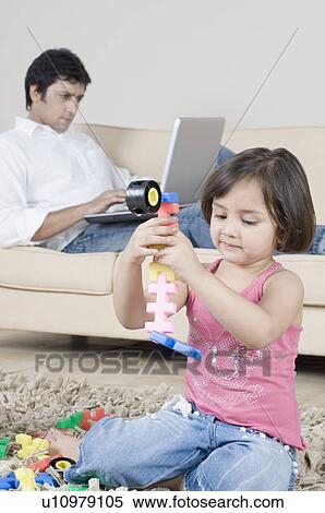Stock Photography - Girl playing with toys and her father working on a laptop in the background. Fotosearch