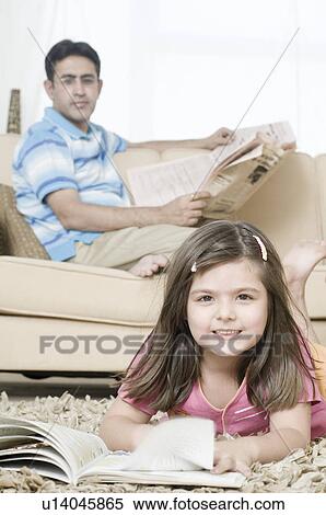 Stock Photography - Portrait of a girl lying in front of a book with her father holding newspaper in the background. Fotosearch