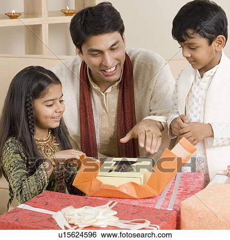Stock Photograph - Young man sitting with his children and opening diwali gifts. Fotosearch