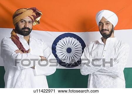 Stock Image - Portrait of a mid adult man and a young man standing in front of an Indian flag with their arms crossed. Fotosearch