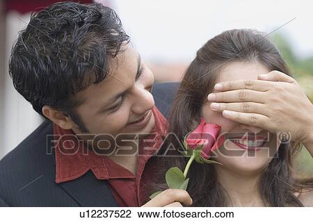Stock Image - Close-up of a young man covering eyes of a young woman and holding a rose. Fotosearch