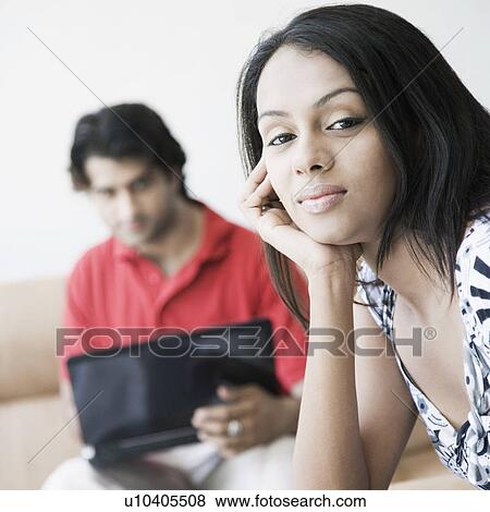 Close-up of a young woman with her hand on her chin and her husband using a laptop in the background View Large Photo Image Stock Photo - Close-up of a young woman with her hand on her chin and her husband using a laptop in the background. Fotosearch