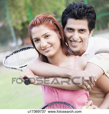 Stock Photo - Portrait of a young man embracing a young woman from behind in a tennis court and smiling. Fotosearch