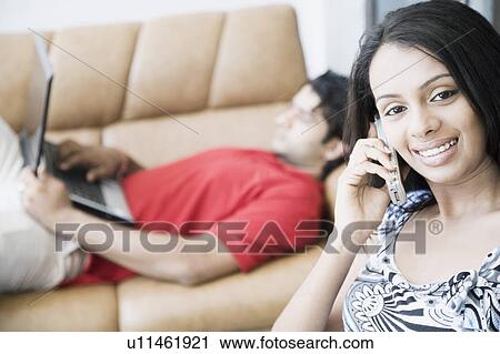 Portrait of a young woman talking on a mobile phone with a young man lying on a couch and using a laptop beside her View Large Photo Image Stock Image - Portrait of a young woman talking on a mobile phone with a young man lying on a couch and using a laptop beside her. Fotosearch
