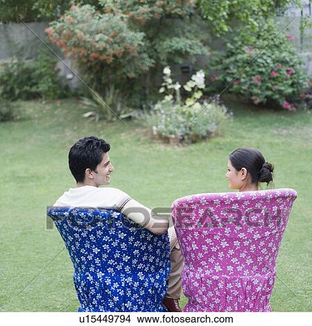 Picture - Rear view of a young couple sitting on chairs in a lawn. Fotosearch