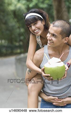 Stock Photo - Portrait of a young couple riding piggyback on a young man and holding a coconut. Fotosearch