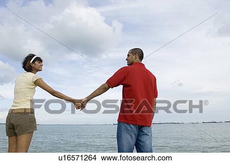 Rear view of a young couple standing on the beach and holding hands View Large Photo Image Picture - Rear view of a young couple standing on the beach and holding hands. Fotosearch