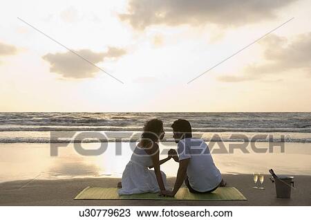 Stock Image - Rear view of a young couple with holding hands sitting on a picnic blanket. Fotosearch