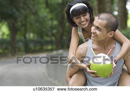 Stock Photo - Young woman riding piggyback on a young man and holding a coconut. Fotosearch