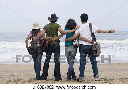 Stock Image - Rear view of two young couples standing on the beach with their arms around each other. Fotosearch
