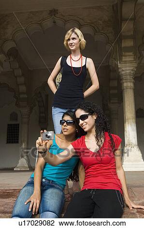Stock Image - Two young women taking a picture with their friend standing behind them, Taj Mahal, Agra, Uttar Pradesh, India. Fotosearch