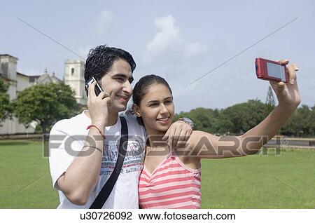 Stock Image - Close-up of a young couple talking on a photograph of themselves with a cathedral in the background, Se Cathedral, Goa, India. Fotosearch