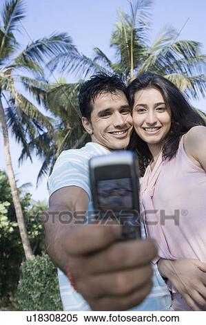 Stock Photography - Low angle view of a young couple taking a picture of themselves with a mobile phone and smiling. Fotosearch